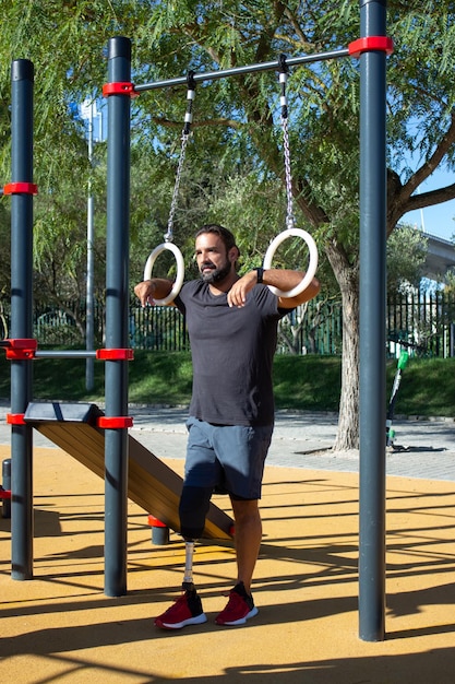 Junger Mann mit Beinprothese mit Turnringen zum Training. Starker Sportler, der auf dem Sportplatz steht und Ringe mit Händen hält, die weit schauen. Gesundheitsversorgung, Sport für Menschen mit Behinderungen