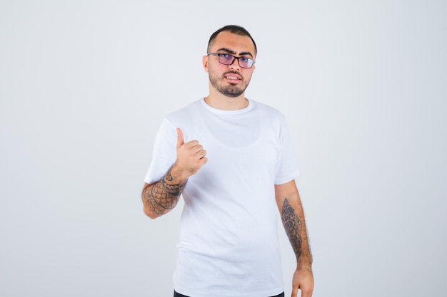 Junger Mann mit Brille und Daumen nach oben in weißem T-Shirt und schwarzer Hose und glücklich aussehend