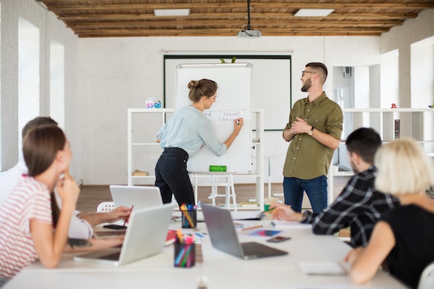 Junger Mann mit Brille und Hemd und hübsche Frau in Bluse, die in der Nähe des Boards stehen und den Kollegen fröhlich ein neues Projekt präsentieren Gruppe kreativer Menschen, die in einem modernen weißen Büro zusammenarbeiten