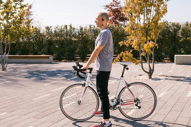 Junger Mann mit dem drahtlosen Bluetooth, das mit weißem Fahrrad im Park steht