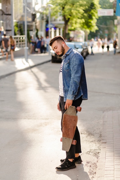 Junger Mann mit dem hölzernen Skateboard, das auf Straße steht