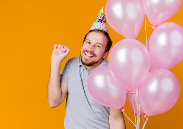 Junger Mann mit Feiertagsmütze, die Geburtstagsfeier feiert, die Bündel Luftballons glücklich und aufgeregt lächelt, die fröhlich über orange Wand stehen