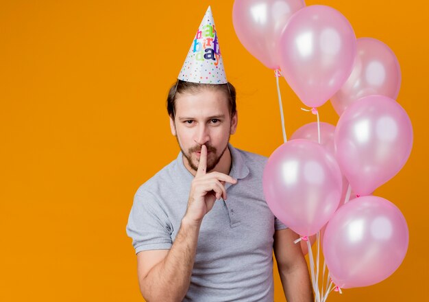 Junger Mann mit Feiertagsmütze, die Geburtstagsfeier feiert, die Bündel von Luftballons hält, die Stille gasture mit Finger auf den Lippen stehen, die über orange Wand stehen