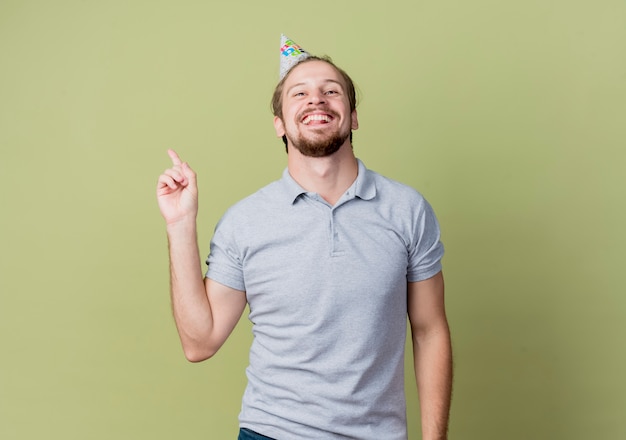 Junger Mann mit Feiertagsmütze, die Geburtstagsfeier glücklich und aufgeregt zeigt Zeigefinger über Lichtwand zeigt