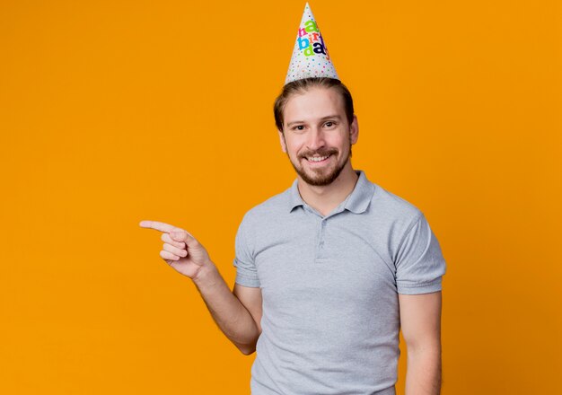 Junger Mann mit Feiertagsmütze feiert Geburtstagsfeier glücklich und fröhlich lächelnd mit dem Finger zur Seite stehend über orange Wand