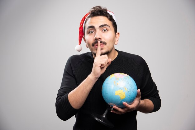 Junger Mann mit Globus in Santas Hut zeigt eine Geste der Stille.