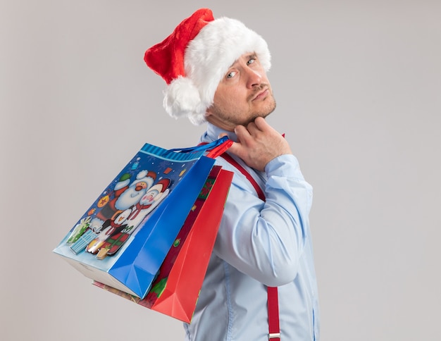 Kostenloses Foto junger mann mit hosenträgerfliege in weihnachtsmütze mit geschenkpapiertüten, die mit traurigem ausdruck auf weißem hintergrund in die kamera schaut