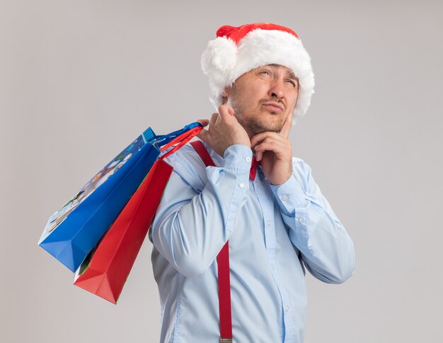 Junger Mann mit Hosenträgerfliege in Weihnachtsmütze mit Geschenkpapiertüten, die verwirrt auf weißem Hintergrund nachschlagen