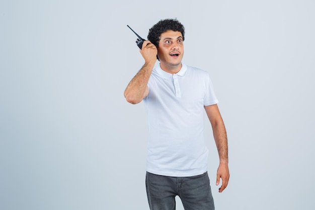 Junger Mann mit Polizei-Walkie-Talkie-Telefon in weißem T-Shirt, Hose und erstauntem Blick. Vorderansicht.