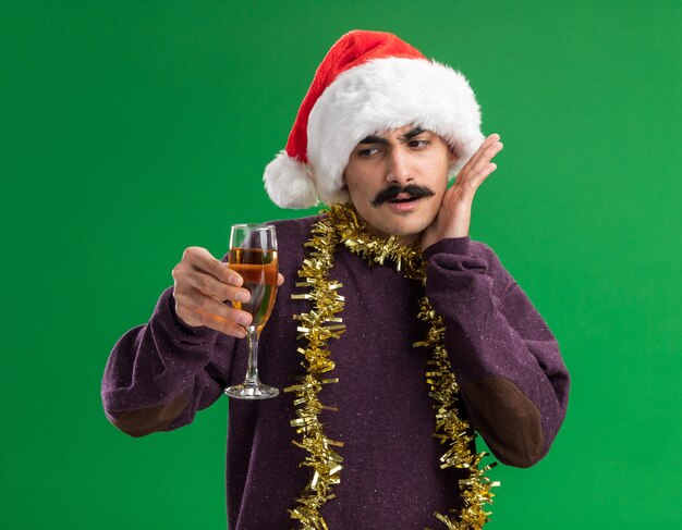 Junger Mann mit Schnurrbart, der Weihnachtsweihnachtsmütze mit Lametta um seinen Hals hält, der Glas Champagner hält, der es verwirrt betrachtet, über grünem Hintergrund stehend