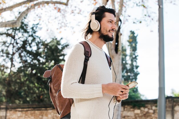 Junger Mann mit seiner hörenden Musik des Rucksacks am Handy durch Kopfhörer an draußen