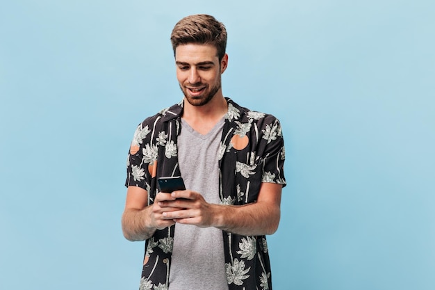 Junger Mann mit trendiger Frisur und Bart in grauem, coolem T-Shirt und sommerlichem, modernem Kurzarmhemd, der ins Telefon schaut und lächelt