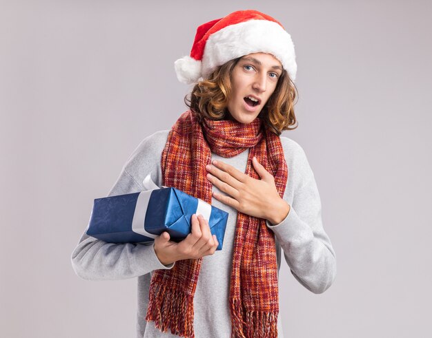 Junger mann mit weihnachtsmütze mit warmem schal um den hals hält weihnachtsgeschenk und sieht überrascht aus und fühlt sich dankbar, über weißer wand zu stehen