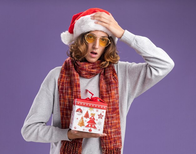 Junger Mann mit Weihnachtsmütze und gelber Brille mit warmem Schal um den Hals, der ein Weihnachtsgeschenk hält, das mit der Hand über dem Kopf über der lila Wand verwechselt wird?