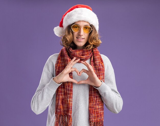 Junger Mann mit Weihnachtsmütze und gelber Brille mit warmem Schal um den Hals lächelt und macht Herzgeste mit Händen und Fingern, die über lila Wand stehen