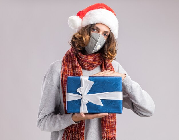 Junger Mann mit Weihnachtsmütze und Gesichtsschutzmaske mit warmem Schal um den Hals, der Weihnachtsgeschenk mit ernstem Gesicht über weißer Wand hält