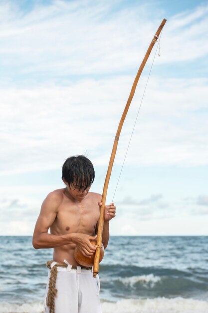 Junger Mann ohne Hemd am Strand mit Holzbogen, der sich darauf vorbereitet, Capoeira zu üben