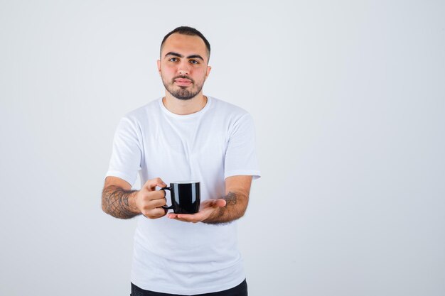 Junger Mann präsentiert eine Tasse Tee in weißem T-Shirt und schwarzer Hose und sieht ernst aus