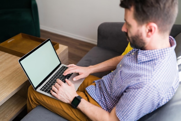 Junger Mann sitzt auf dem Sofa und tippt auf seinem Laptop mit leerem Bildschirm und Kopierbereich