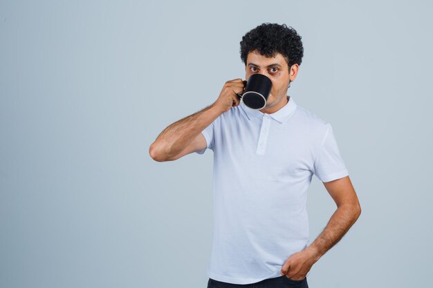 Junger Mann trinkt eine Tasse Tee, während er die Hand in weißem T-Shirt und Jeans an der Taille hält und ernst aussieht. Vorderansicht.