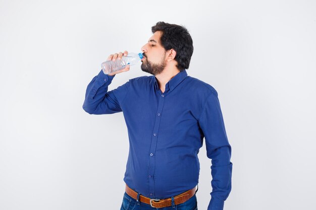Junger Mann trinkt Wasser in blauem Hemd und Jeans und sieht ernst aus, Vorderansicht.
