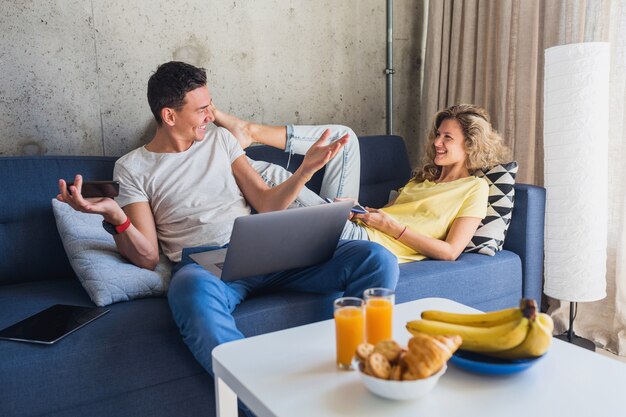 Junger Mann und Frau bleiben allein zu Hause sitzen auf Sofa mit Geräten, die online arbeiten