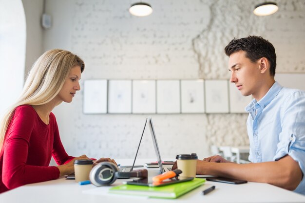 Junger Mann und Frau sitzen am Tisch von Angesicht zu Angesicht und arbeiten am Laptop im mitarbeitenden Büro