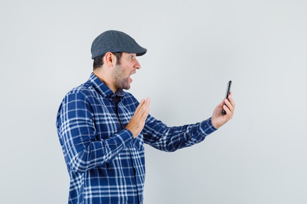 Junger Mann winkt Hand auf Video-Chat in Hemd, Mütze und schaut fröhlich. Vorderansicht.