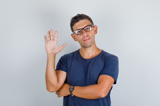 Junger Mann winkt Hand in dunkelblauem T-Shirt, Brille und schaut glücklich, Vorderansicht.