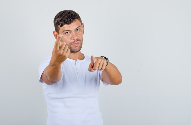 Junger Mann winkt und zeigt Finger auf Kamera im weißen T-Shirt und schaut positiv, Vorderansicht.