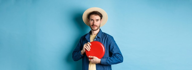 Junger Mann wirft Frisbee und spielt im Sommerurlaub mit Strohhut auf blauem Hintergrund