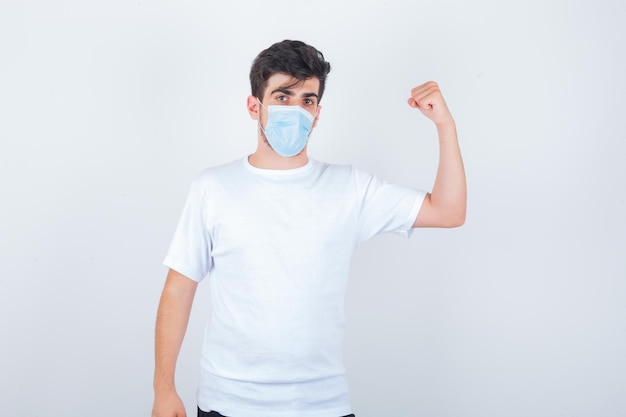 Junger Mann zeigt Armmuskeln in weißem T-Shirt, Maske und sieht mächtig aus looking