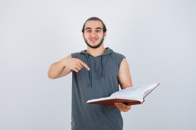 Junger Mann zeigt auf Buch im ärmellosen Kapuzenpulli und sieht hübsch aus, Vorderansicht.