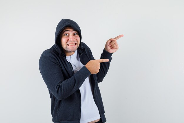 Junger Mann zeigt auf die rechte Seite in T-Shirt, Jacke und sieht fröhlich aus. Vorderansicht.