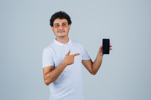 Junger Mann zeigt auf Handy im weißen T-Shirt und sieht selbstbewusst aus. Vorderansicht.