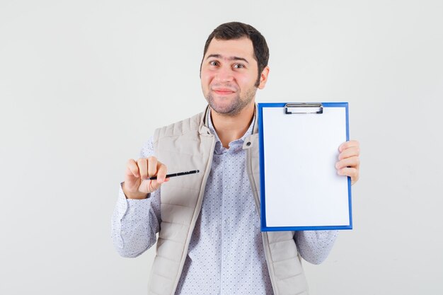 Junger Mann zeigt auf Notizbuch mit Stift in beiger Jacke und schaut optimistisch, Vorderansicht.