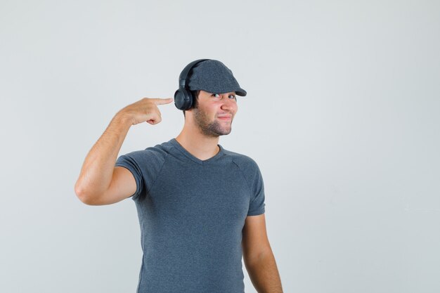 Junger Mann zeigt auf seine Kopfhörer in T-Shirt, Mütze und sieht fröhlich aus. Vorderansicht.