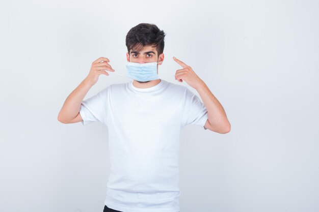 Junger Mann zeigt auf seinen Kopf in weißem T-Shirt, Maske und sieht selbstbewusst aus looking