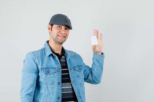 Junger Mann zeigt Flasche Pillen in Jacke, Mütze und schaut froh, Vorderansicht.