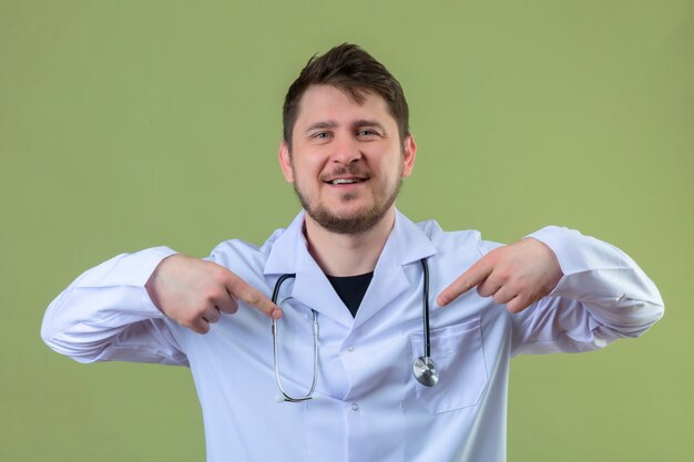 Junger Mannarzt, der weißen Kittel und Stethoskop trägt, das mit Lächeln auf Gesicht zuversichtlich schaut und sich mit den Fingern stolz und glücklich über isoliertem grünem Hintergrund zeigt
