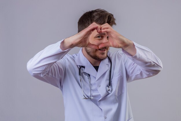 Junger Mannarzt, der weißen Kittel und Stethoskop trägt, die Herzform mit Hand und Fingern lächelnd durch das Zeichen schauend tun
