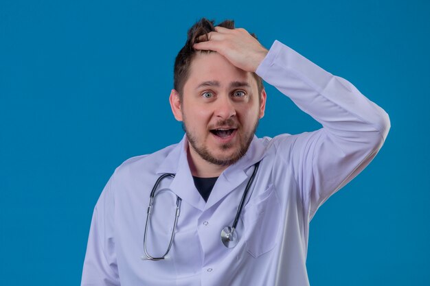 Junger Mannarzt, der weißen Kittel und Stethoskop trägt, überrascht mit glücklichem Gesicht über lokalisiertem blauem Hintergrund