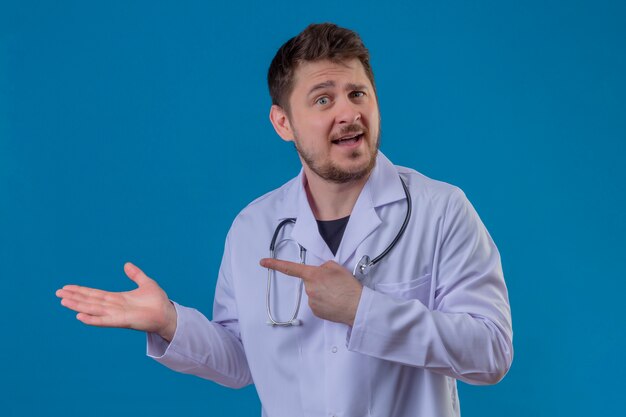 Junger Mannarzt, der weißen Kittel und Stethoskop trägt, zeigt mit Hand und Finger zur Seite über lokalisiertem blauem Hintergrund