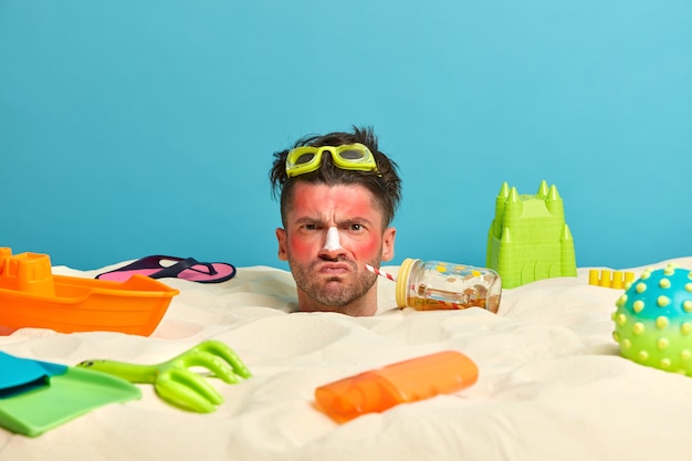 Junger Mannkopf mit Sonnenschutzcreme auf Gesicht umgeben von Strandaccessoires