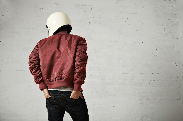 Junger Motorradfahrer in weißem Helm und roter Nylonjacke schoss von hinten mit den Händen in den Gesäßtaschen seiner Jeans in einem Studio mit weißen Wänden.