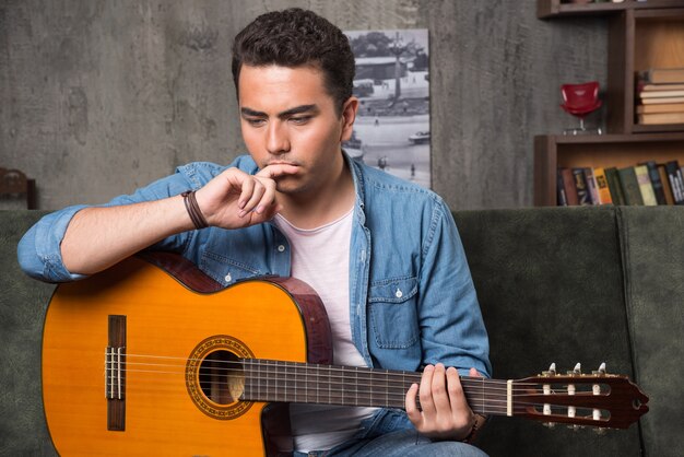 Junger Musiker, der eine schöne Gitarre hält und auf Sofa sitzt. Hochwertiges Foto