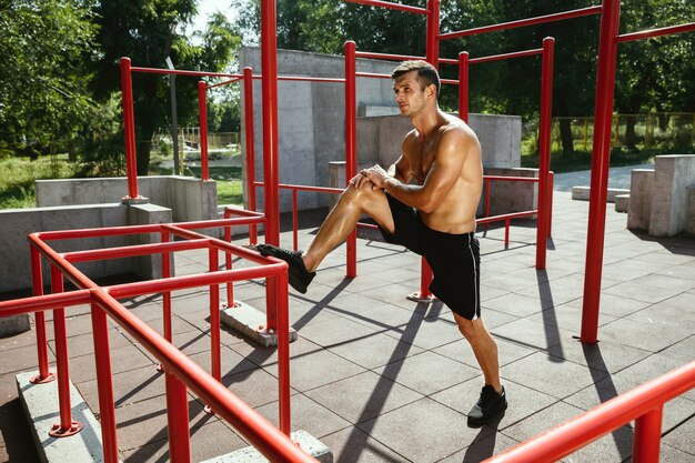 Junger muskulöser hemdloser kaukasischer Mann, der Streckübungen am Spielplatz am sonnigen Sommertag tut. Trainiere seinen Oberkörper im Freien. Konzept von Sport, Training, gesundem Lebensstil, Wohlbefinden.