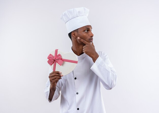 Junger nachdenklicher afroamerikanischer Koch in der Kochuniform hält herzförmige Schachtel und legt Hand auf Kinn lokalisiert auf weißer Wand