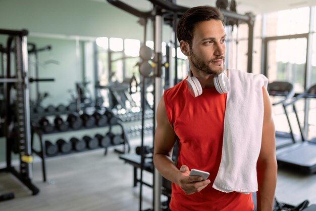 Junger nachdenklicher Athlet, der Smartphone in einem Fitnessstudio verwendet