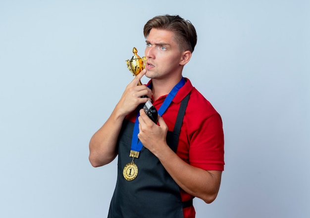 Junger nachdenklicher blonder männlicher Friseur in Uniform mit Goldmedaille hält Siegerpokal und Haarschneider, die Seite lokalisiert auf weißem Raum mit Kopienraum betrachten